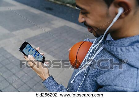 Stock Image - Black man holding basketball listening to cell phone with earbuds. Fotosearch