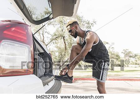 Stock Photography - Black man leaning on car hatchback tying shoe. Fotosearch
