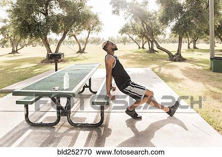 Stock Image - Black man leaning on picnic table in park stretching leg. Fotosearch