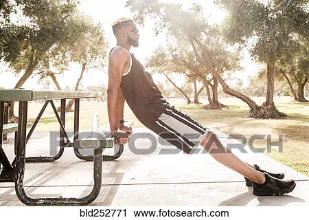 Stock Image - Black man leaning on picnic table in park stretching legs. Fotosearch