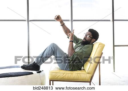 Stock Image - Black man sitting in chair posing for cell phone selfie. Fotosearch