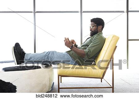 Stock Image - Black man sitting in chair texting on cell phone. Fotosearch