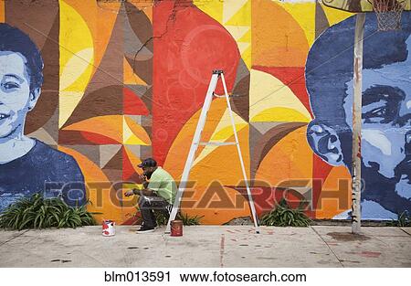 Stock Image - Black man sitting on ladder by mural. Fotosearch