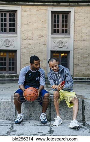 Picture - Black men sitting on concrete with basketball and texting on cell phone. Fotosearch