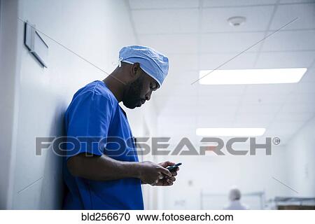 Stock Image - Black nurse leaning on wall texting on cell phone. Fotosearch