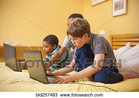 Stock Image - Boys sitting on bed using laptops. Fotosearch