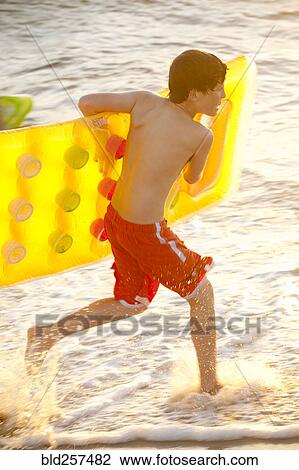 Stock Image - Caucasian boy running into ocean carrying inflatable raft. Fotosearch