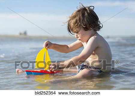 Picture - Caucasian boy sitting in ocean playing with toy sailboat. Fotosearch