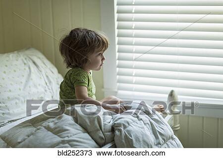 Stock Image - Caucasian boy sitting on bed near window. Fotosearch