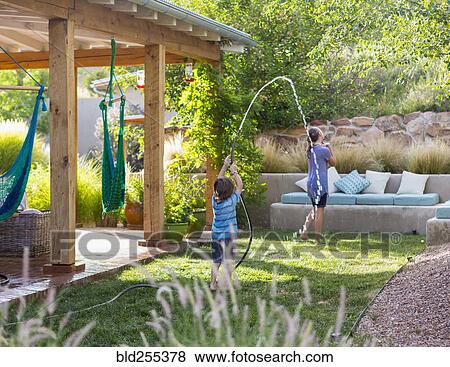 Stock Photo - Caucasian boy spraying sister with hose in backyard. Fotosearch
