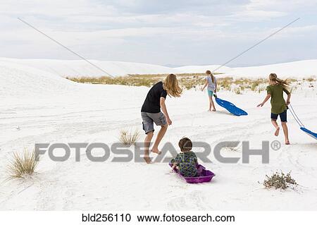 Stock Image - Caucasian boys and girls playing on sleds in sand. Fotosearch