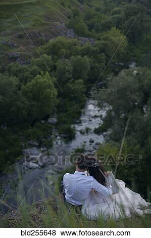 Stock Photo - Caucasian bride and groom sitting on hill admiring river. Fotosearch