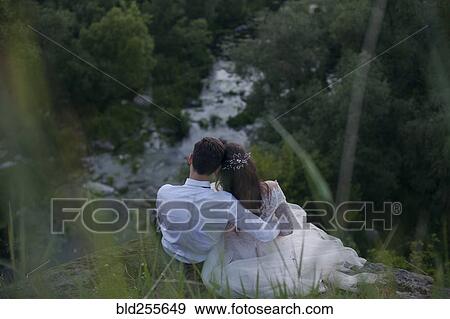 Stock Photo - Caucasian bride and groom sitting on hill admiring river. Fotosearch