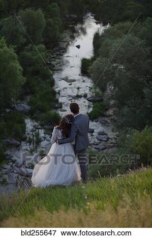 Stock Photo - Caucasian bride and groom standing on hill admiring river. Fotosearch