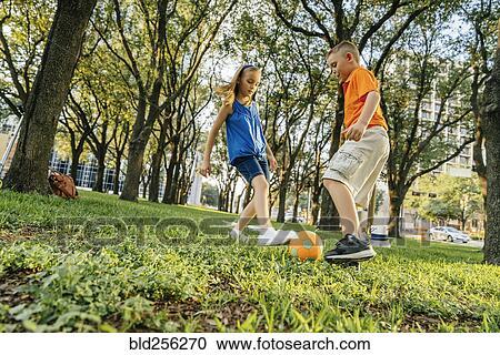 Stock Image - Caucasian brother and sister kicking soccer ball in park. Fotosearch