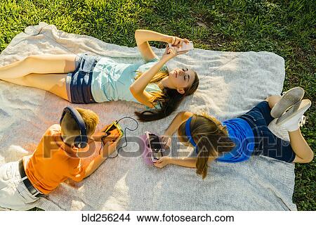 Caucasian brother and sisters laying on blanket in park using technology View Large Photo Image Picture - Caucasian brother and sisters laying on blanket in park using technology. Fotosearch