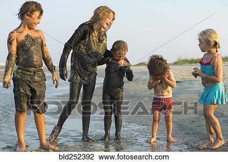 Stock Image - Caucasian brothers and sisters covered in mud playing on beach. Fotosearch