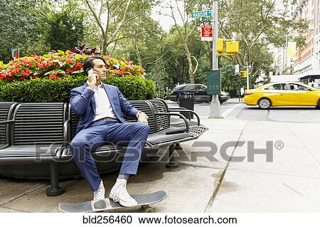 Stock Image - Caucasian businessman sitting on bench in city talking on cell phone. Fotosearch
