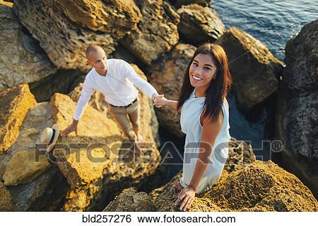Stock Photograph - Caucasian couple climbing on rocks at beach. Fotosearch