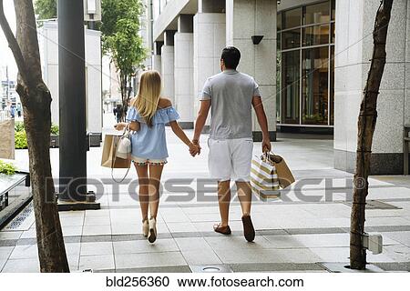 Stock Image - Caucasian couple holding hands carrying shopping bags in city. Fotosearch