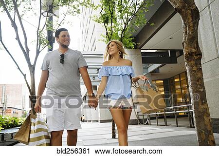 Stock Image - Caucasian couple holding hands carrying shopping bags in city. Fotosearch