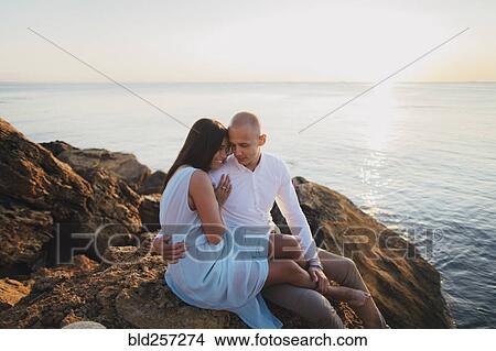 Picture - Caucasian couple hugging on rocks on beach at sunset. Fotosearch