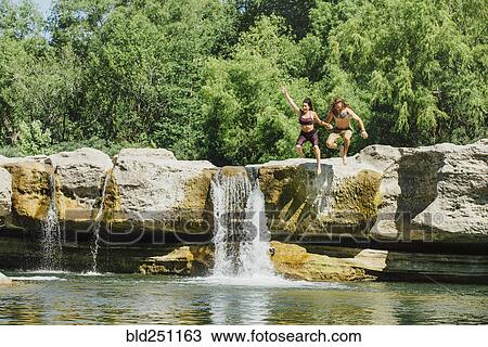 Stock Image - Caucasian couple jumping off rocks near waterfalls. Fotosearch
