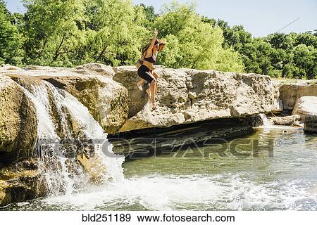 Stock Photo - Caucasian couple jumping off rocks near waterfalls. Fotosearch