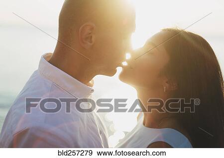 Stock Photo - Caucasian couple kissing near ocean at sunset. Fotosearch