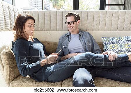Stock Image - Caucasian couple relaxing on sofa texting on cell phone. Fotosearch