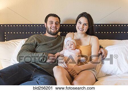 Stock Image - Caucasian couple sitting on bed holding baby daughter. Fotosearch