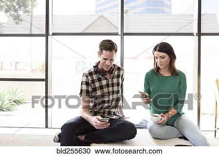 Stock Image - Caucasian couple sitting on bench texting on cell phones. Fotosearch