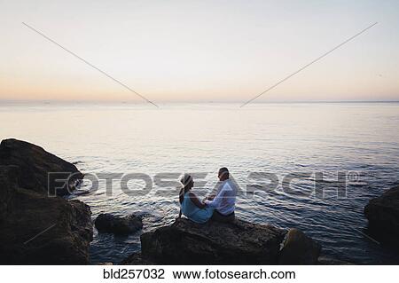 Stock Image - Caucasian couple sitting on rock near ocean at sunset. Fotosearch
