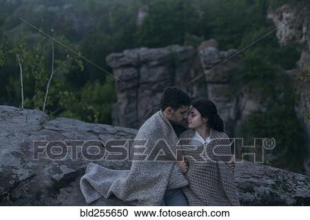 Stock Image - Caucasian couple sitting on rock wrapped in blanket. Fotosearch