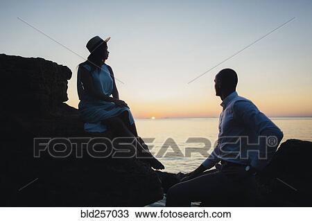 Stock Image - Caucasian couple sitting on rocks near ocean at sunset. Fotosearch