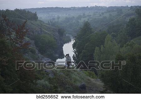 Stock Image - Caucasian couple standing on hill above river. Fotosearch