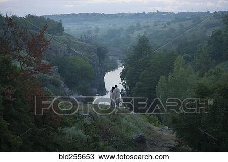 Stock Image - Caucasian couple standing on hill admiring river. Fotosearch