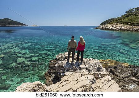 Stock Image - Caucasian couple standing on rock admiring lake. Fotosearch