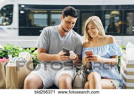 Stock Image - Caucasian couple with shopping bags texting on cell phones near train. Fotosearch