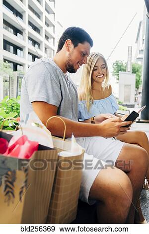 Stock Photo - Caucasian couple with shopping bags texting on cell phones. Fotosearch