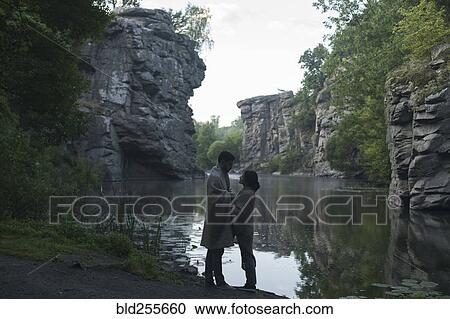 Stock Image - Caucasian couple wrapped in blanket holding hands near river. Fotosearch