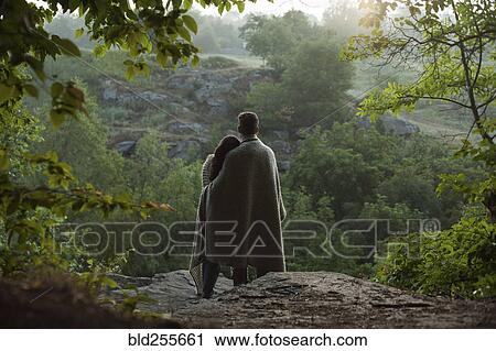 Stock Image - Caucasian couple wrapped in blanket standing on rock in forest. Fotosearch