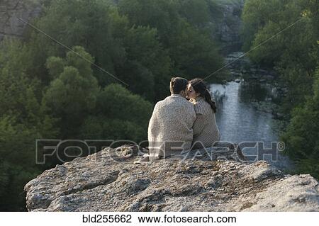 Stock Image - Caucasian couple wrapped in blanket kissing on rock near river. Fotosearch