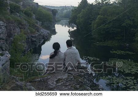 Stock Photo - Caucasian couple wrapped in blanket sitting on rock admiring river. Fotosearch