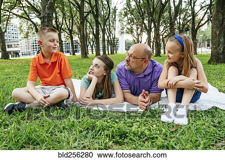 Stock Image - Caucasian father and daughters laying on blanket in park listening to boy. Fotosearch