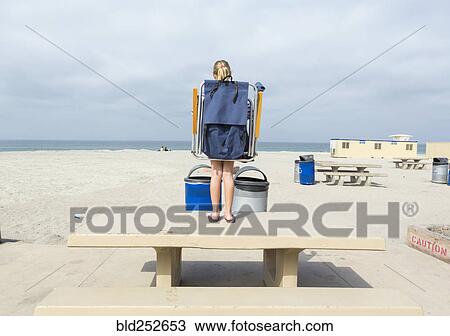 Stock Image - Caucasian girl carrying folding chair on picnic table at beach. Fotosearch