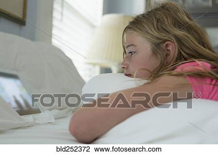 Stock Image - Caucasian girl laying on bed watching digital tablet. Fotosearch