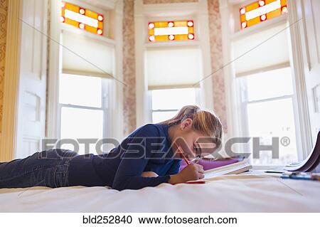 Stock Image - Caucasian girl laying on bed writing in notebook. Fotosearch