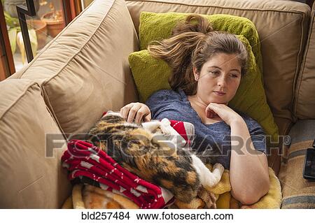 Picture - Caucasian girl laying on sofa petting cat. Fotosearch