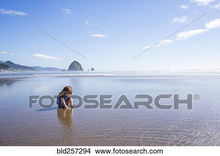 Picture - Caucasian girl sitting on beach in ocean waves. Fotosearch